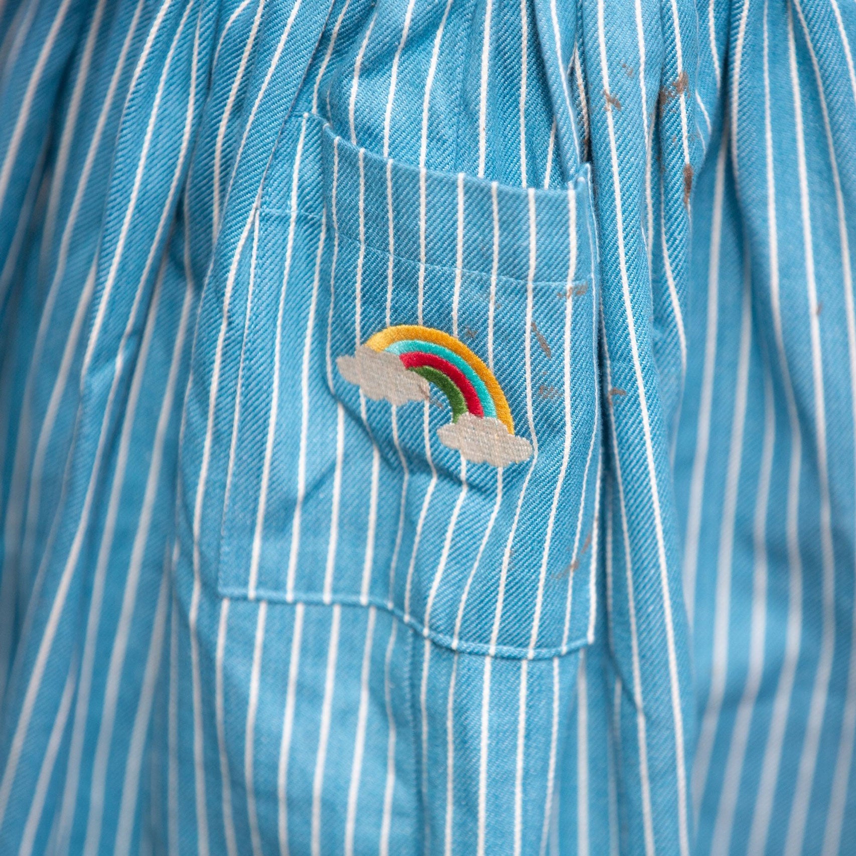 A close up of the rainbow embroidery on the pocket of the Little Green Radicals Child's Organic Cotton Horizon Blue Striped Pinafore Button Dress