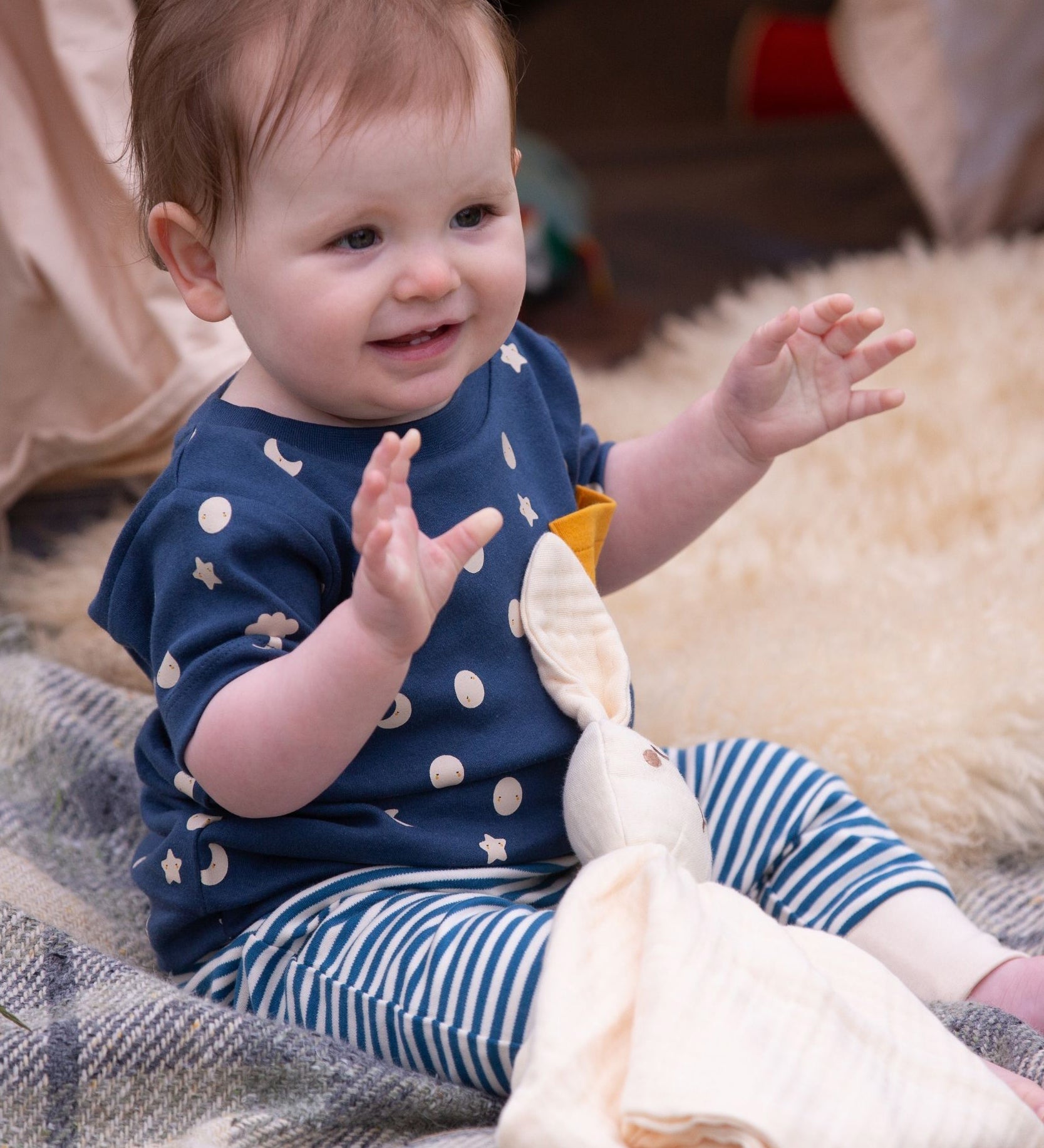 A baby wearing the Little Green Radicals  Full Moon T-Shirt & Jogger outfit set. The baby is sitting on a blanket with a cream coloured bunny comforter toy on their lap.  