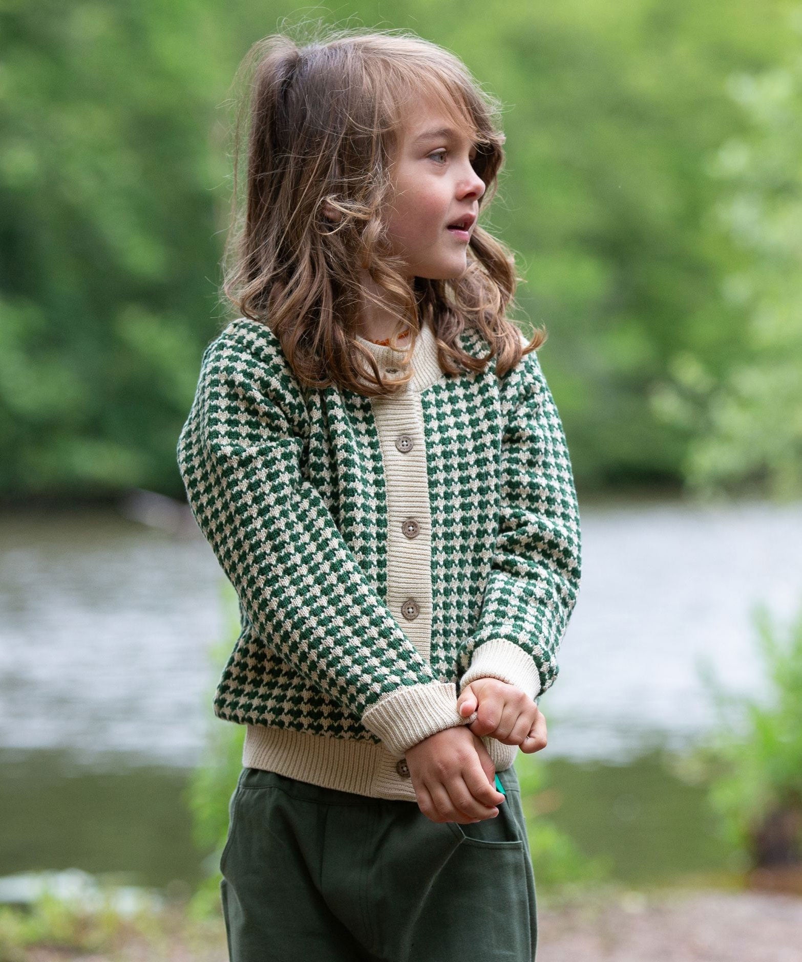 A child wearing the Little Green Radicals From One To Another Olive Green Striped Knitted Child's Organic Cotton Cardigan with green coloured shorts.