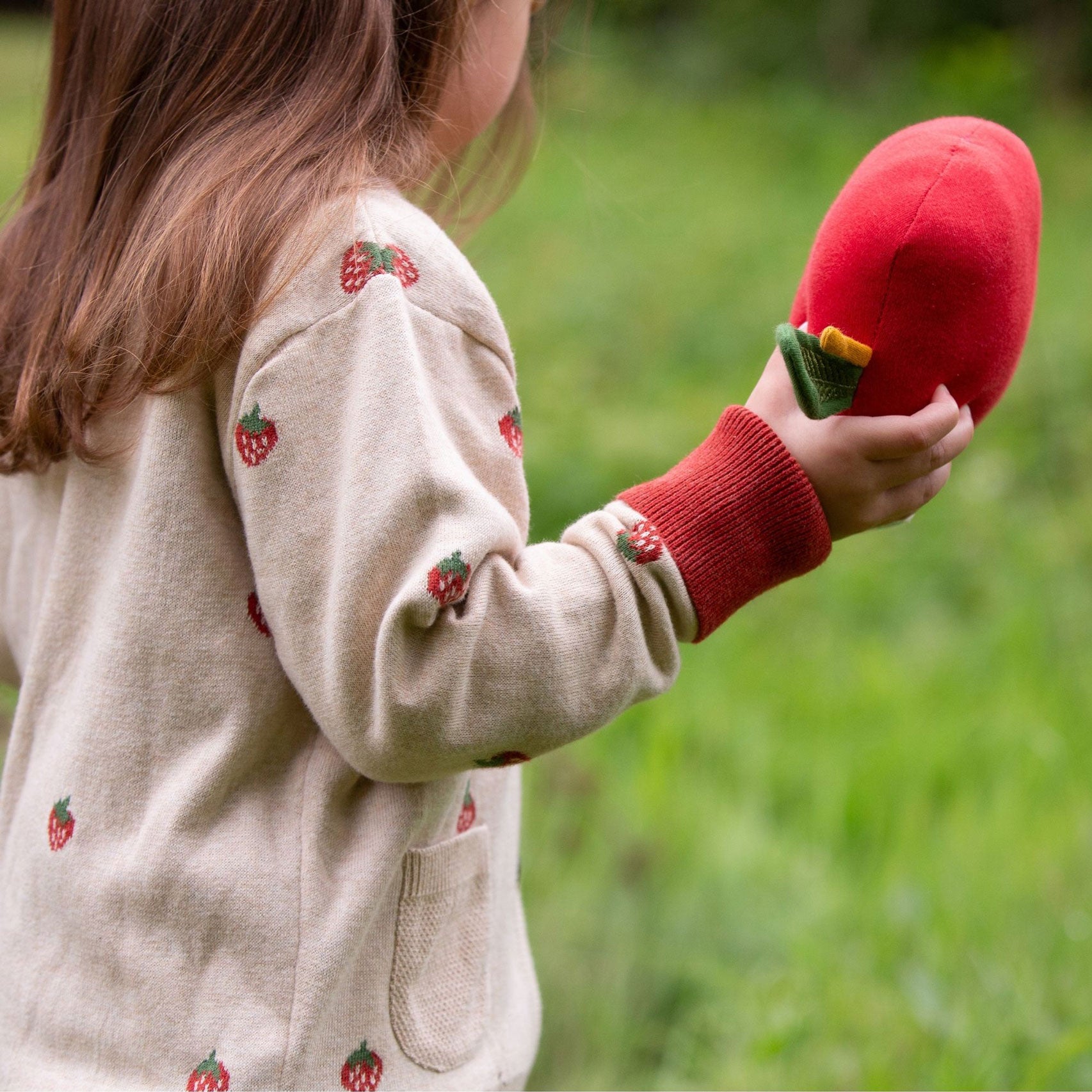 A close up of a child wearing the Little Green Radicals organic From One To Another Strawberry Days print child's Knitted Cardigan. The child is facing away from the camera showing the side of the cardigan