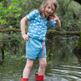 A child wearing the Little Green Radicals Kids Organic Cotton Blue Clouds Loose Fit Short Sleeve Henley Top with a pair of striped blue shorts. The child is stood in water with a pair of red wellies. 