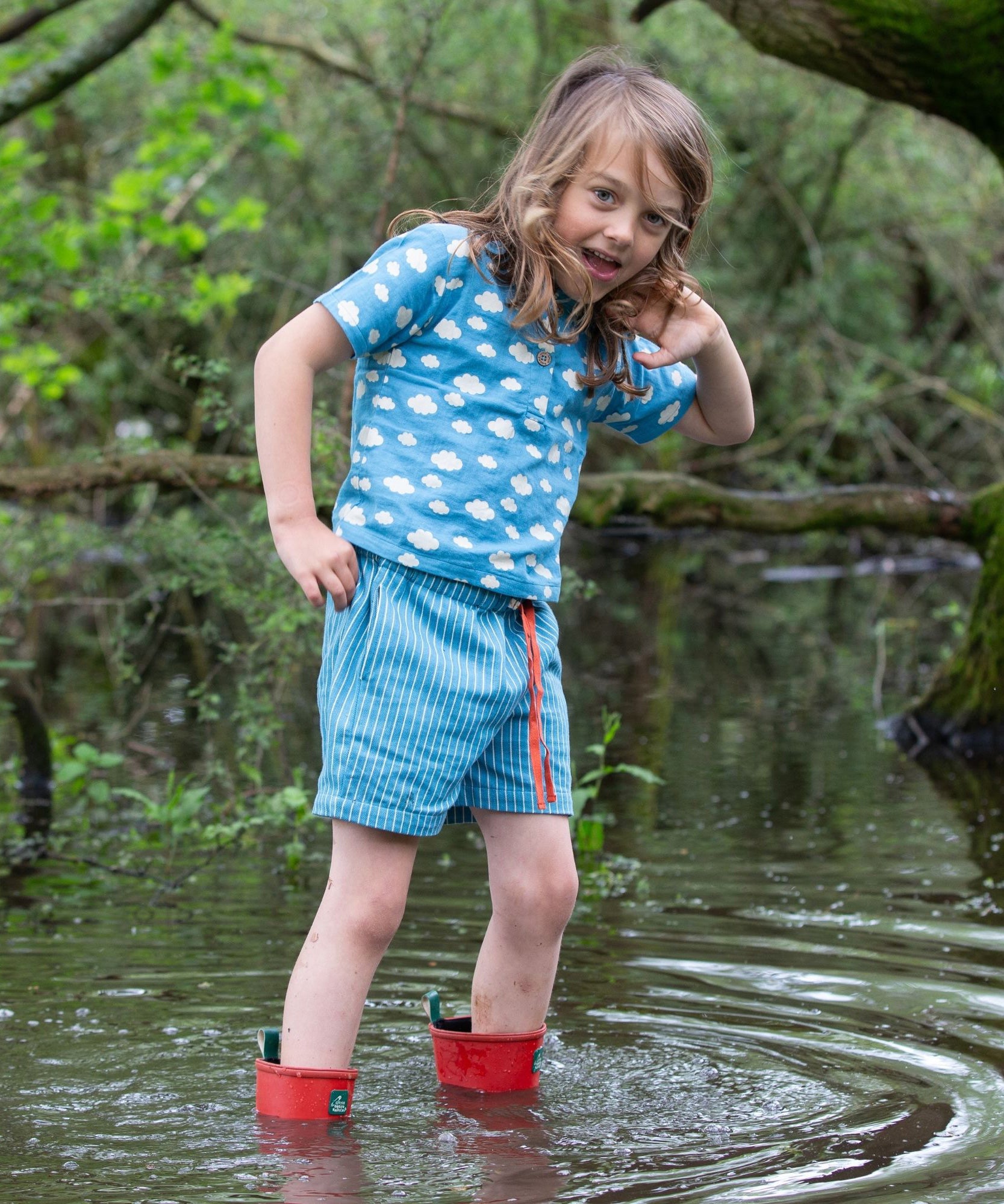 A child wearing the Little Green Radicals Kids Organic Cotton Blue Clouds Loose Fit Short Sleeve Henley Top with a pair of striped blue shorts. The child is stood in water with a pair of red wellies. 