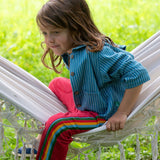 A child wearing the Little Green Radicals Red rainbow stripe Organic Cotton Comfy Joggers with a blue striped shirt. The child is sitting on a white hammock.