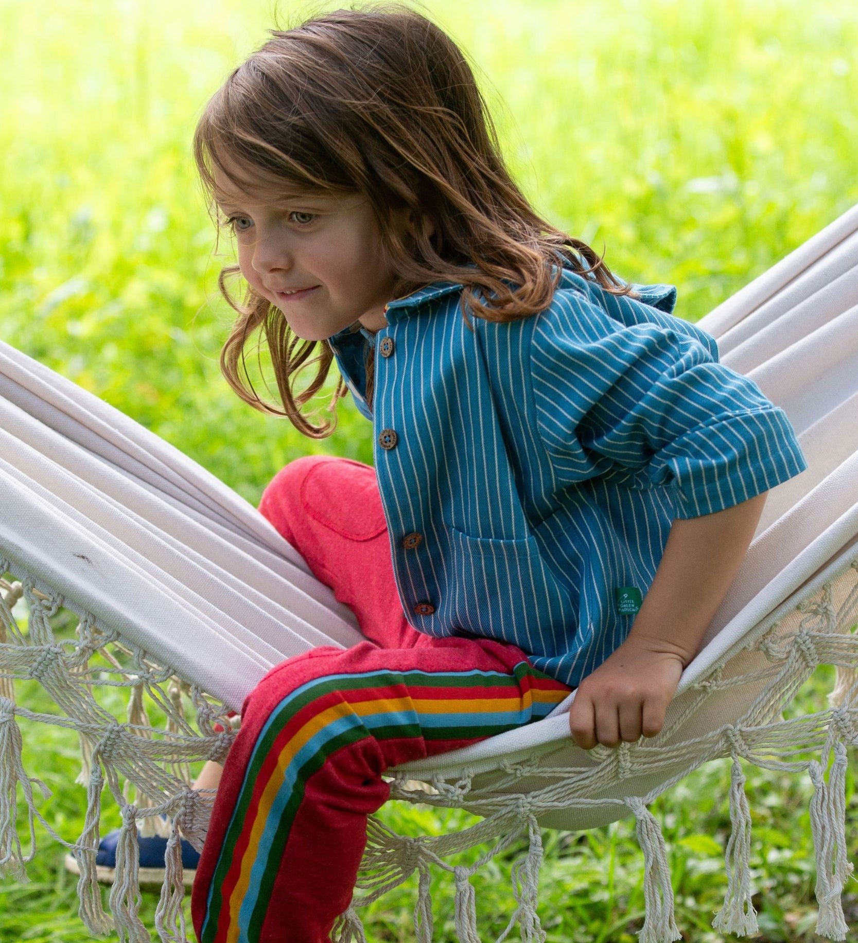 A child wearing the Little Green Radicals Red rainbow stripe Organic Cotton Comfy Joggers with a blue striped shirt. The child is sitting on a white hammock.