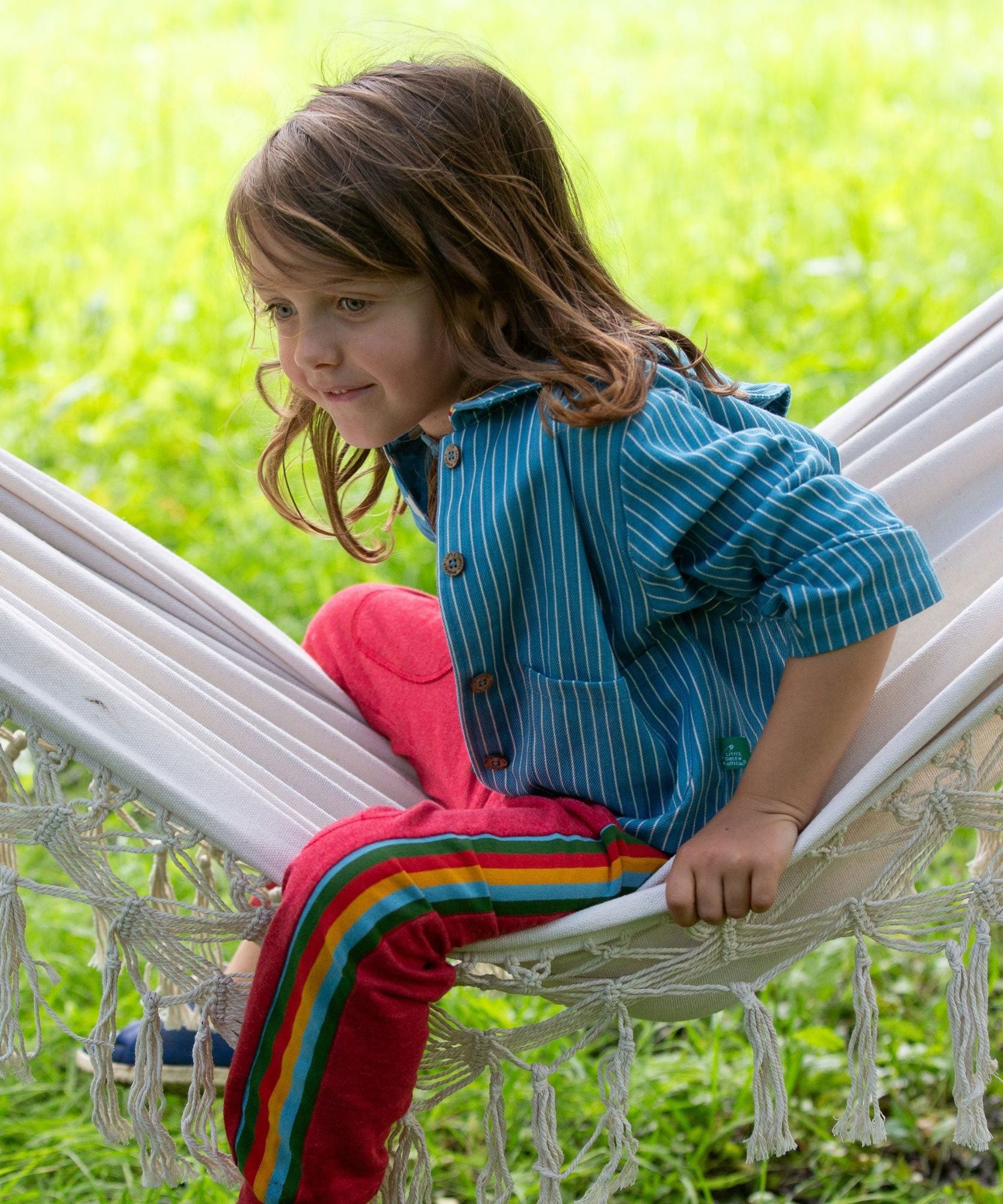 A child wearing the Little Green Radicals Red rainbow stripe Organic Cotton Comfy Joggers with a blue striped shirt. The child is sitting on a white hammock.