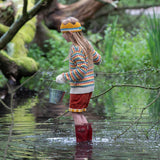 A child wearing the Little Green Radicals From One To Another Rainbow Days Organic Cotton Knitted Cardigan with red coloured shorts. The child is standing in water with red wellington boots and a metal bucket in their hand. 