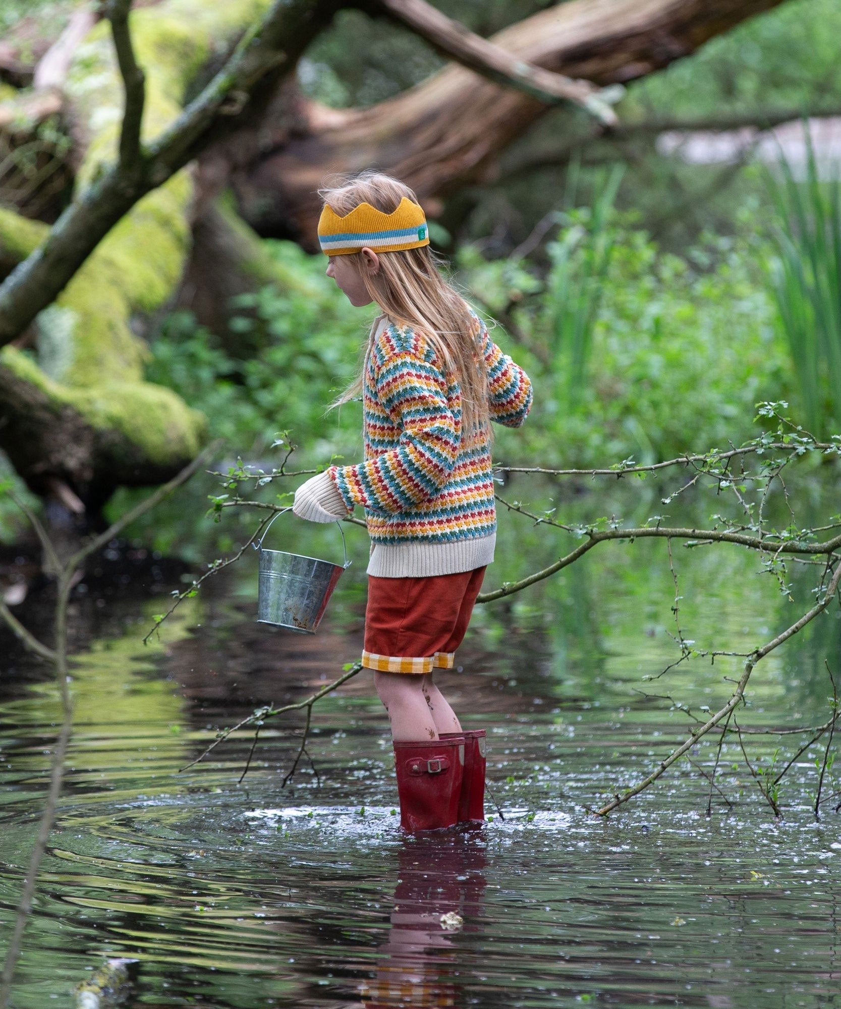 A child wearing the Little Green Radicals From One To Another Rainbow Days Organic Cotton Knitted Cardigan with red coloured shorts. The child is standing in water with red wellington boots and a metal bucket in their hand. 