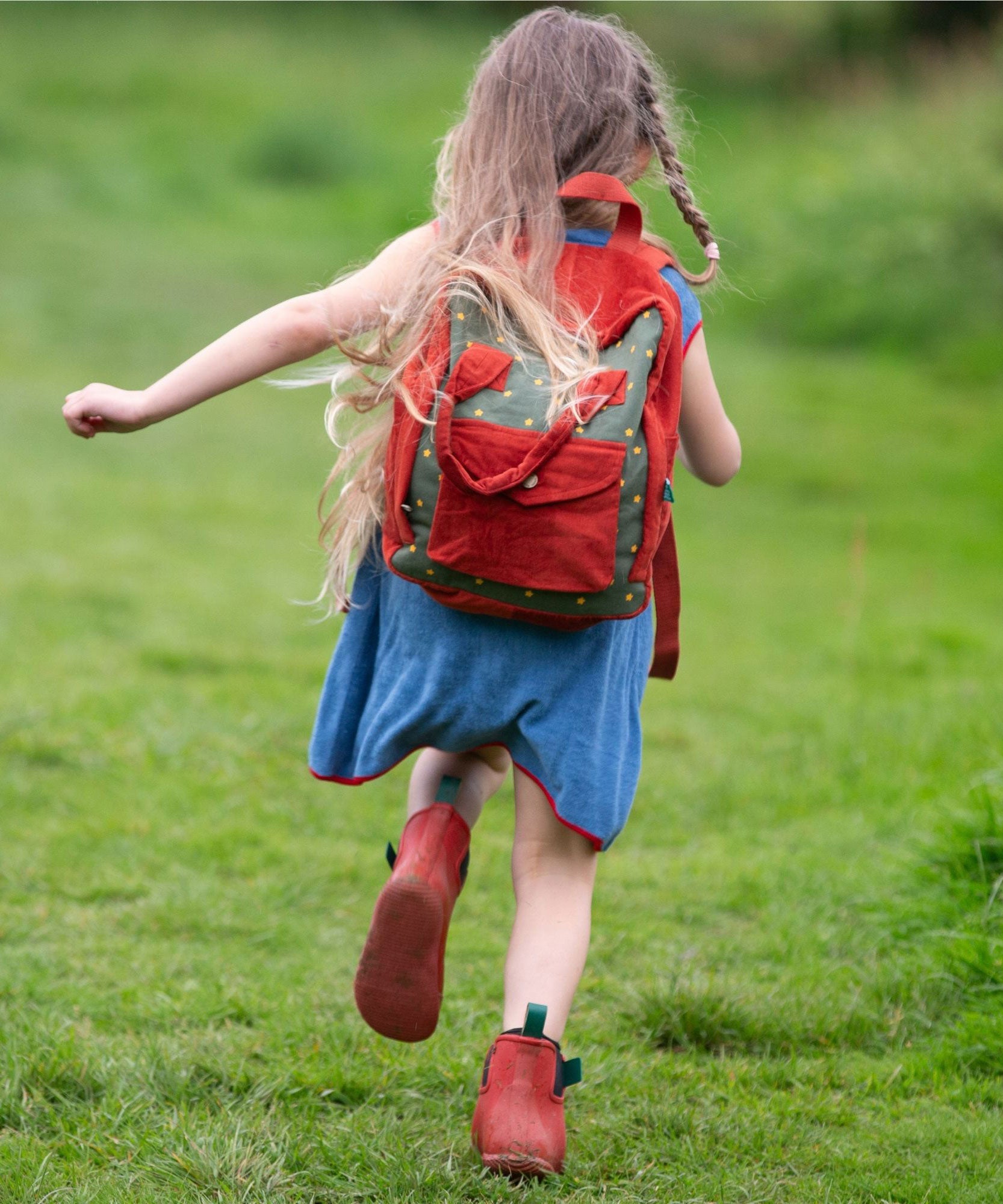 A child running wearing the Little Green Radicals Kids Ready For Adventure Yellow Stars print red and green Backpack on their back. The child is facing away from the camera giving a full view of the backpack