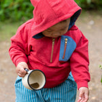 A child wearing the Little Green Radicals Child's Red Organic Quarter Zip Hoodie with blue striped shorts. The child has an enamel mug in their hand with muddy water inside.