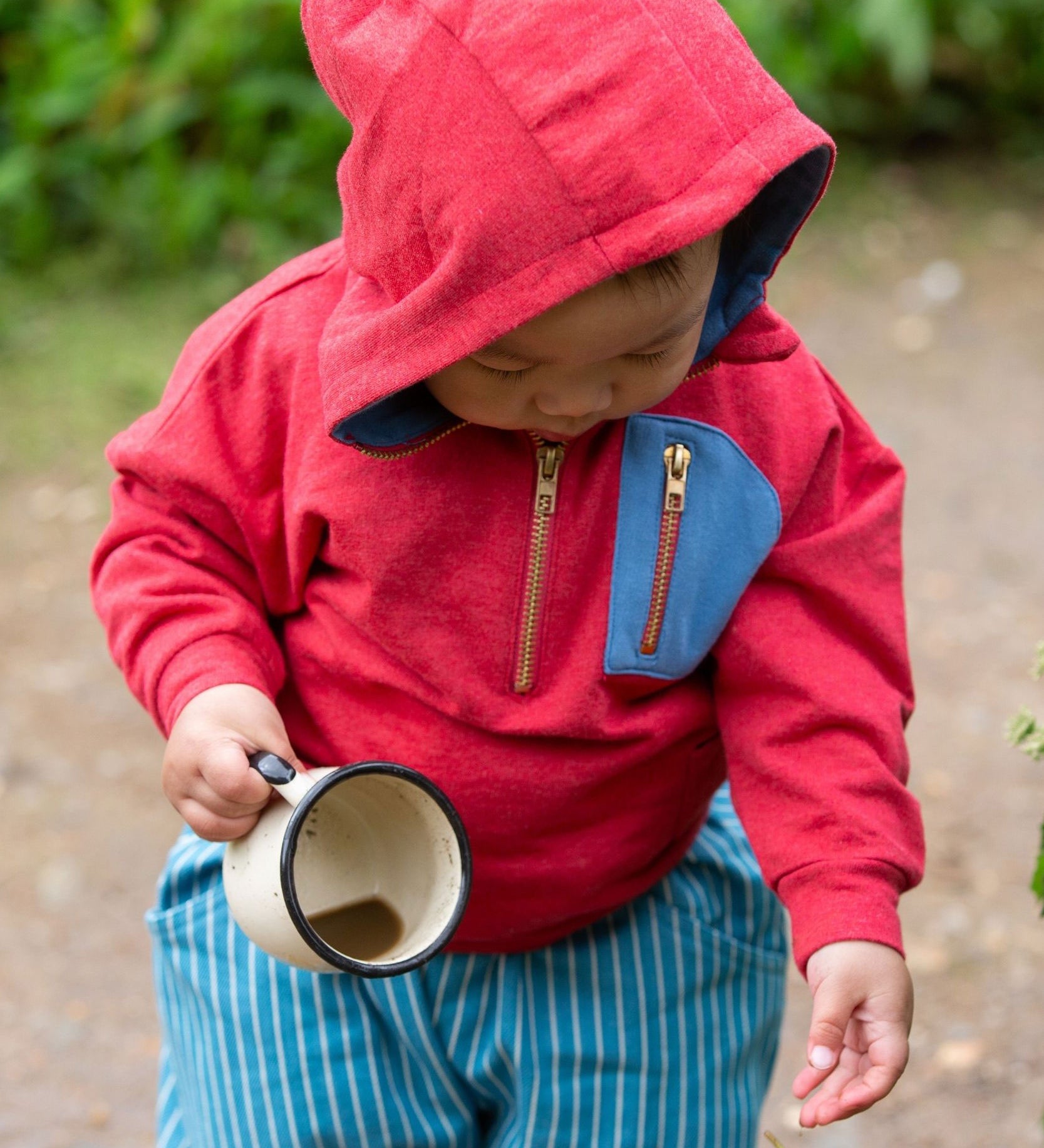 A child wearing the Little Green Radicals Child's Red Organic Quarter Zip Hoodie with blue striped shorts. The child has an enamel mug in their hand with muddy water inside.