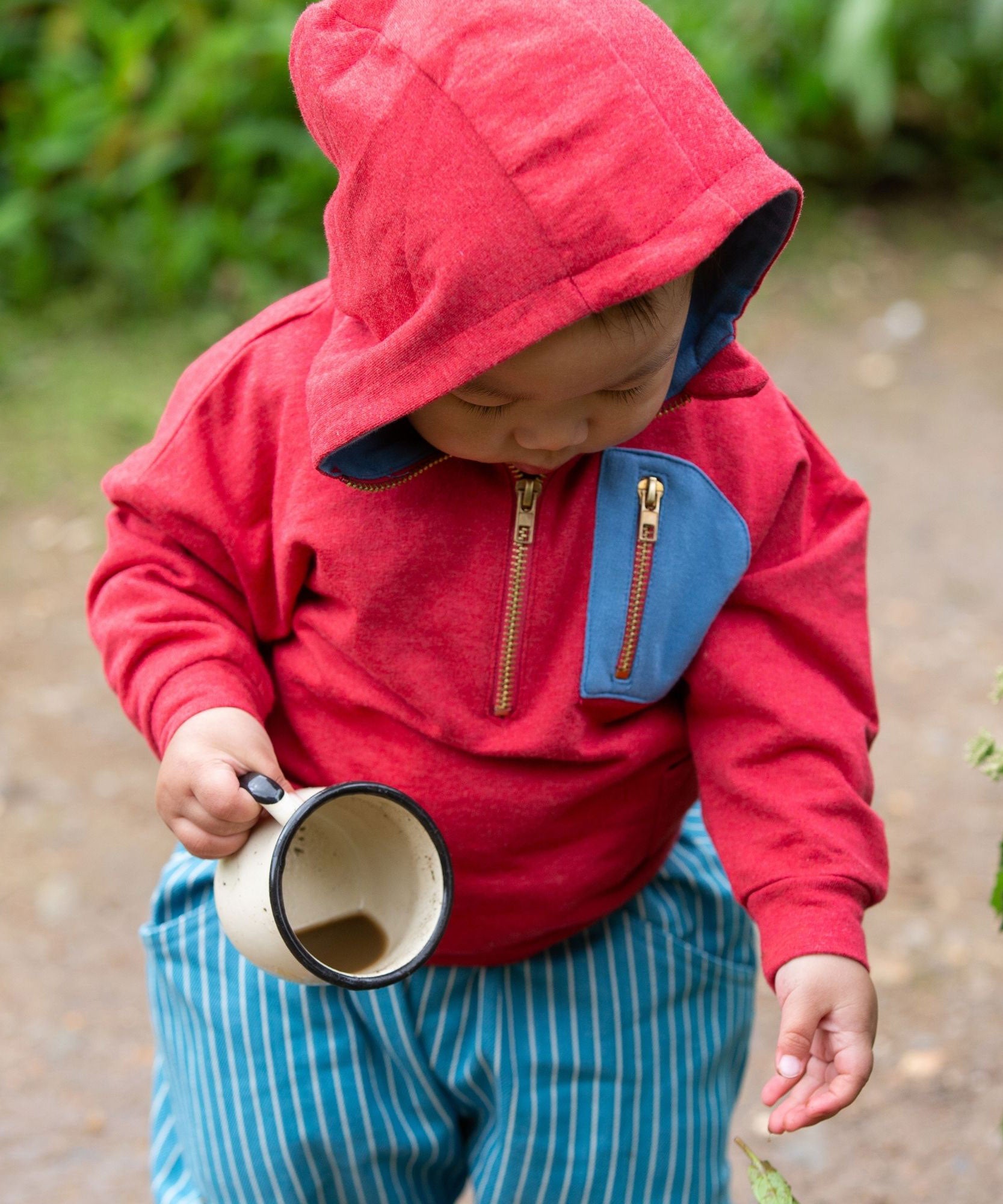 A child wearing the Little Green Radicals Child's Red Organic Quarter Zip Hoodie with blue striped shorts. The child has an enamel mug in their hand with muddy water inside.