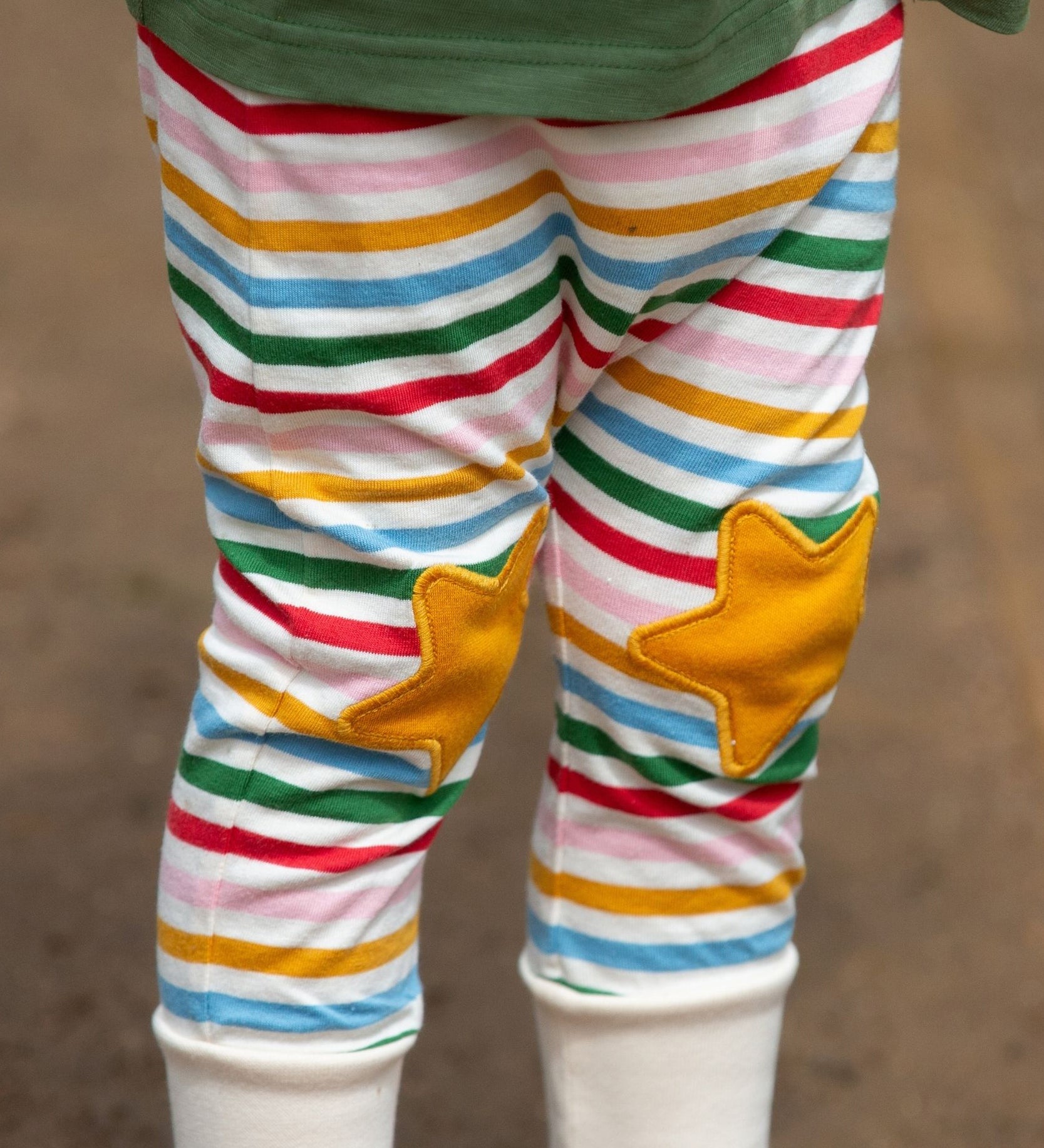 A close up of a child's legs wearing the Little Green Radicals Stars Knee Patch Rainbow Striped Children's Organic Cotton Joggers