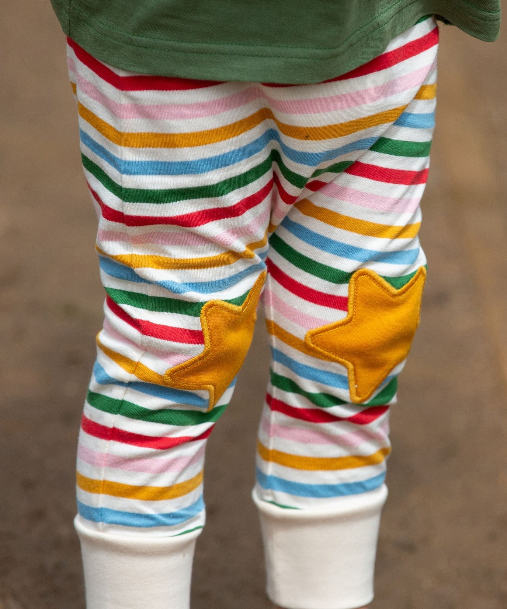 A close up of a child's legs wearing the Little Green Radicals Stars Knee Patch Rainbow Striped Children's Organic Cotton Joggers