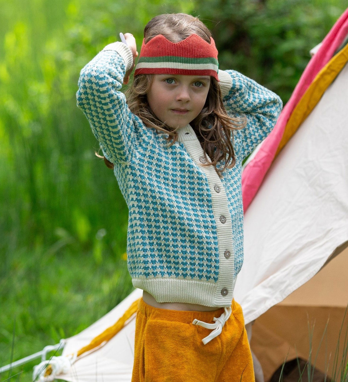 A child wearing the blue and white From One To Another Knitted Cardigan by Little Green Radicals with yellow gold coloured shorts and a knitted red crown. 