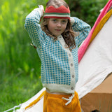 A child wearing the blue and white From One To Another Knitted Cardigan by Little Green Radicals with yellow gold coloured shorts and a knitted red crown. 