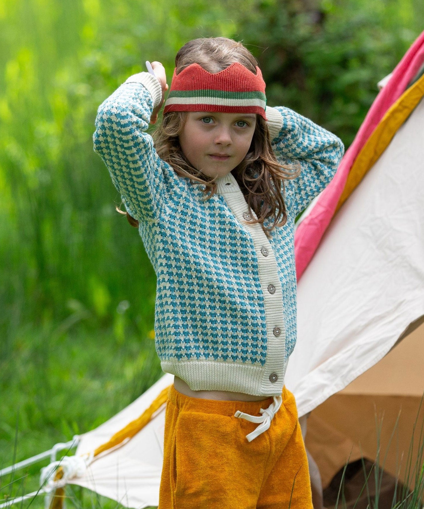 A child wearing the blue and white From One To Another Knitted Cardigan by Little Green Radicals with yellow gold coloured shorts and a knitted red crown. 