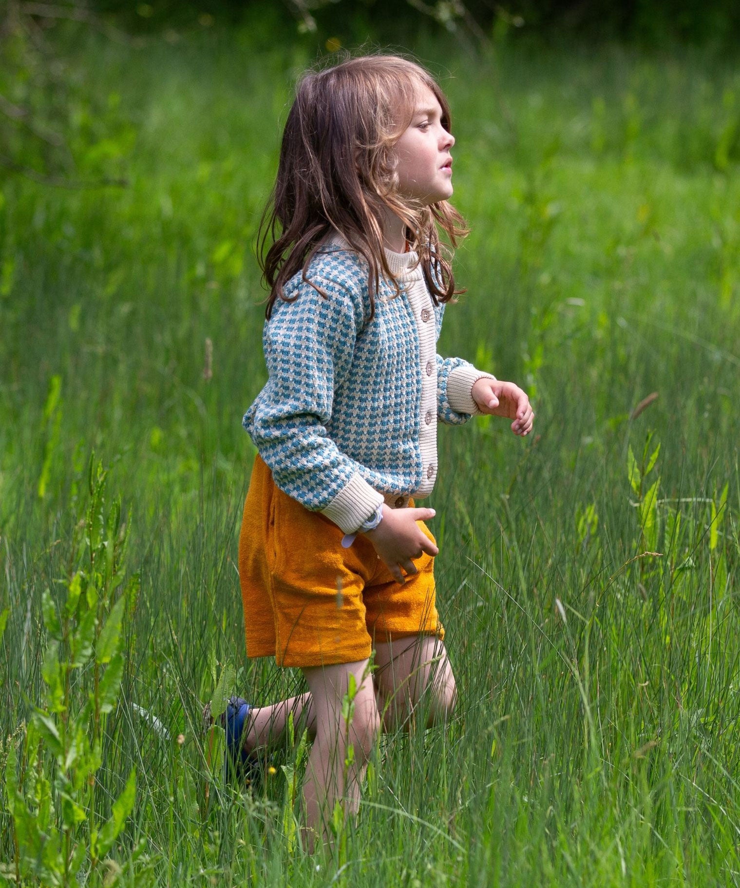 A child wearing the blue and white From One To Another Knitted Cardigan by Little Green Radicals with yellow gold coloured shorts. The child is running through long green grass.