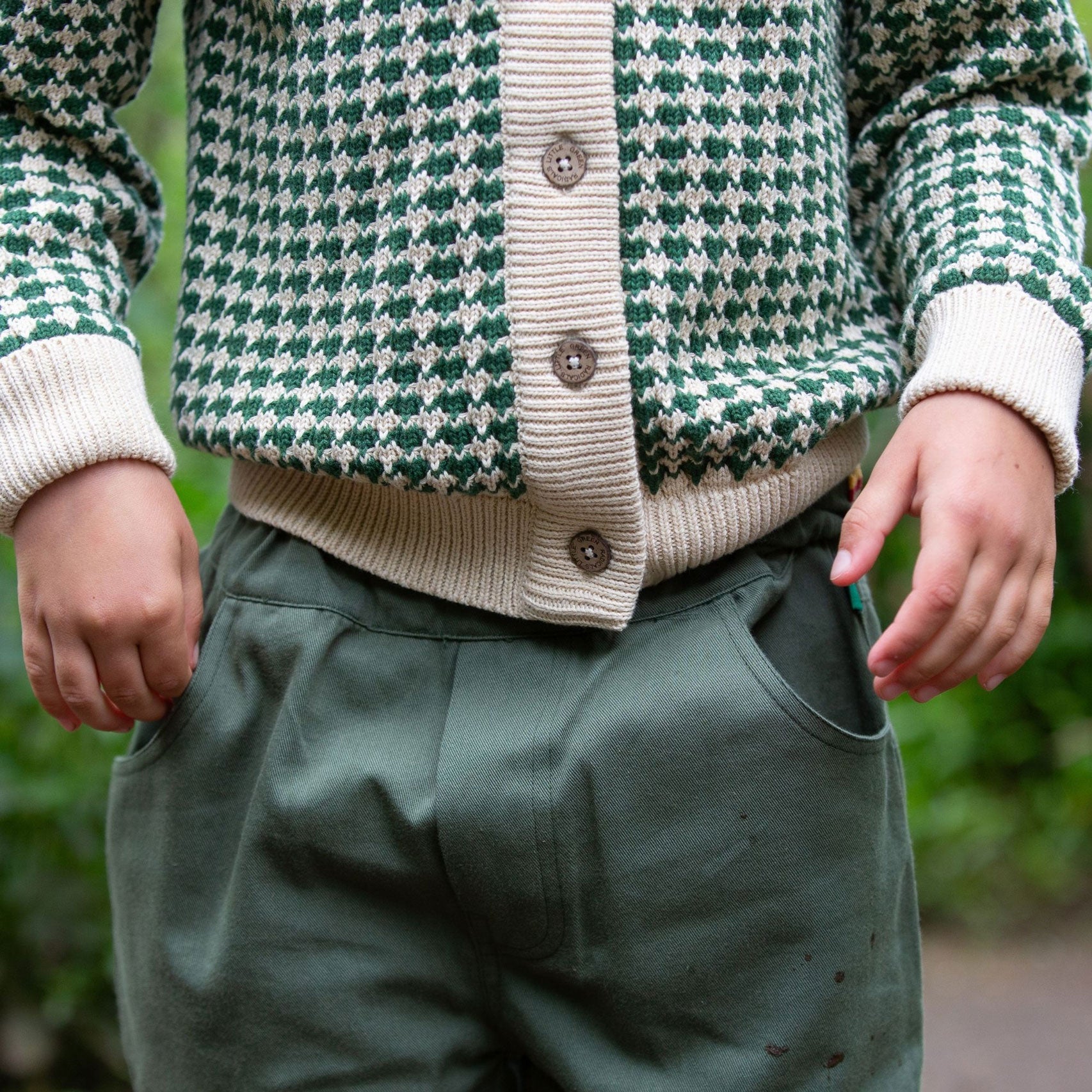 A close up of a child wearing the Little Green Radicals From One To Another Olive Green Striped Knitted Child's Organic Cotton Cardigan with green coloured shorts. The branded buttons can be seen on the cardigan