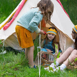 A child wearing the blue and white From One To Another Knitted Cardigan by Little Green Radicals with yellow gold coloured shorts. The child is standing outside a bell tent. Two other children can be seen sitting on the grass in front of them.