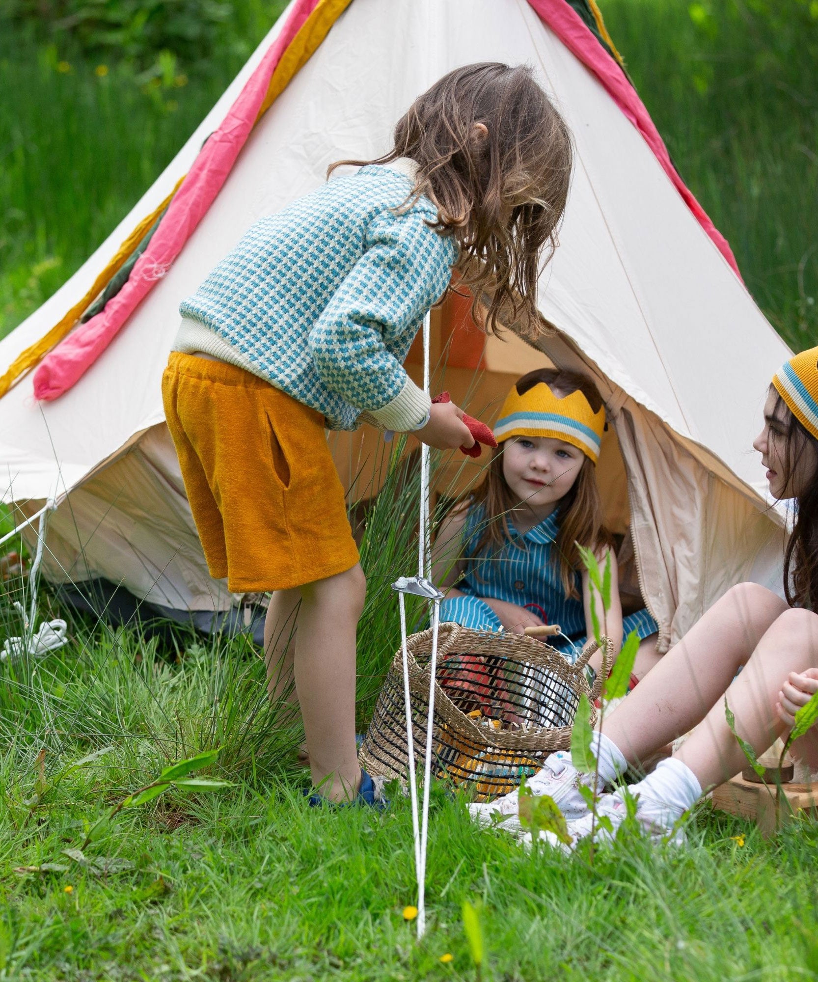 A child wearing the blue and white From One To Another Knitted Cardigan by Little Green Radicals with yellow gold coloured shorts. The child is standing outside a bell tent. Two other children can be seen sitting on the grass in front of them.