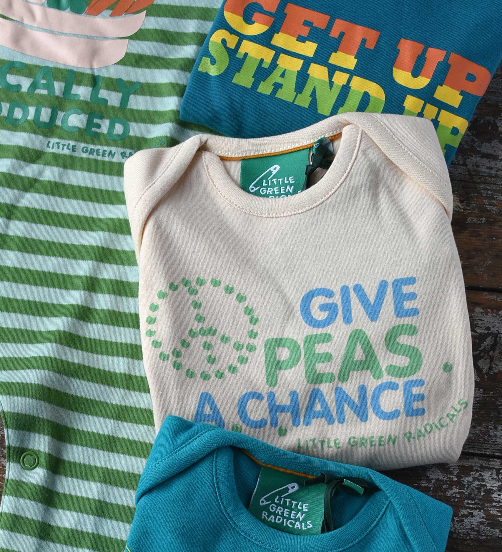 A close up of the give peas a chance slogan Little Green Radicals playsuit folded up. The other three slogan playsuits can be seen placed around it.