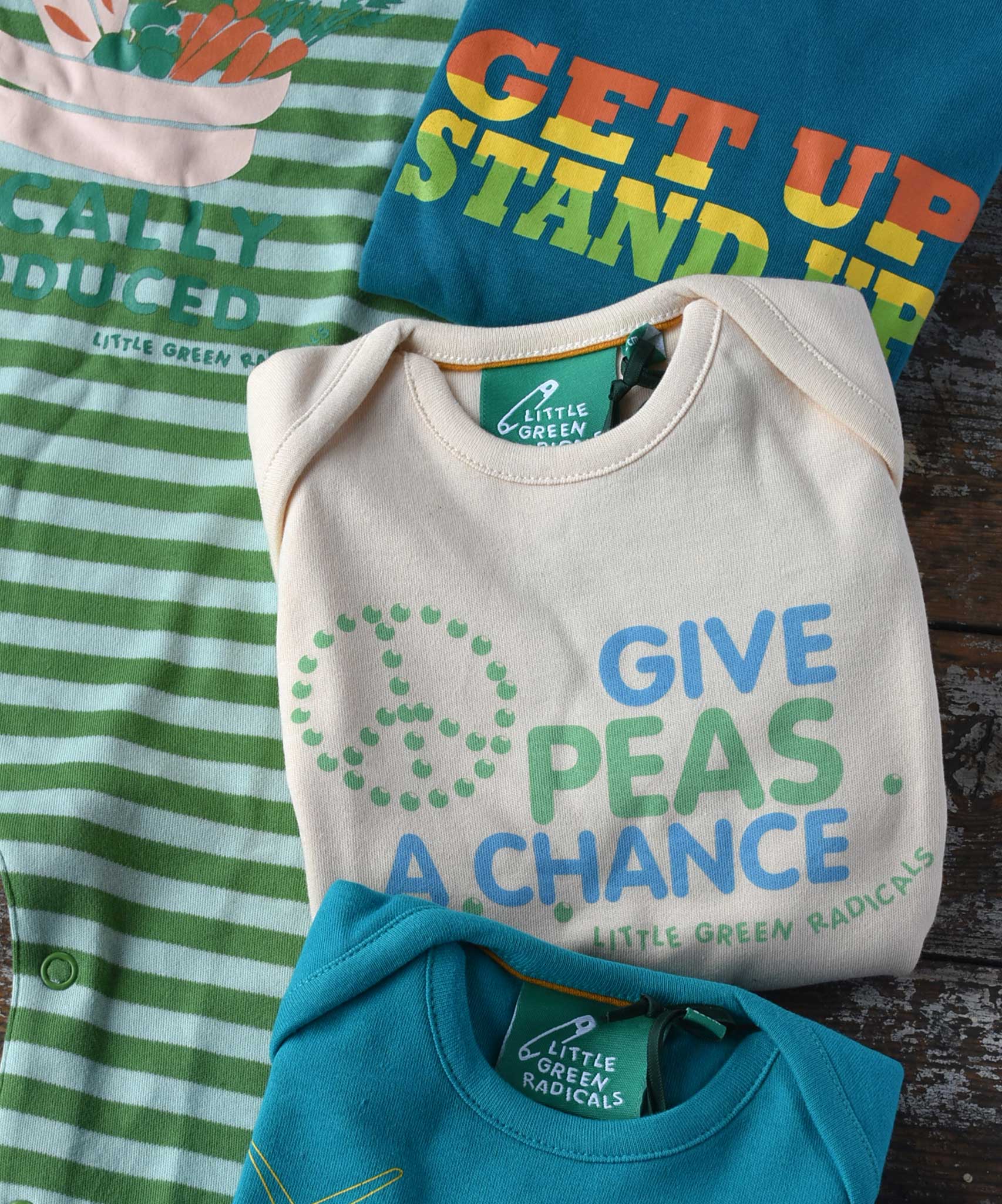 A close up of the give peas a chance slogan Little Green Radicals playsuit folded up. The other three slogan playsuits can be seen placed around it.