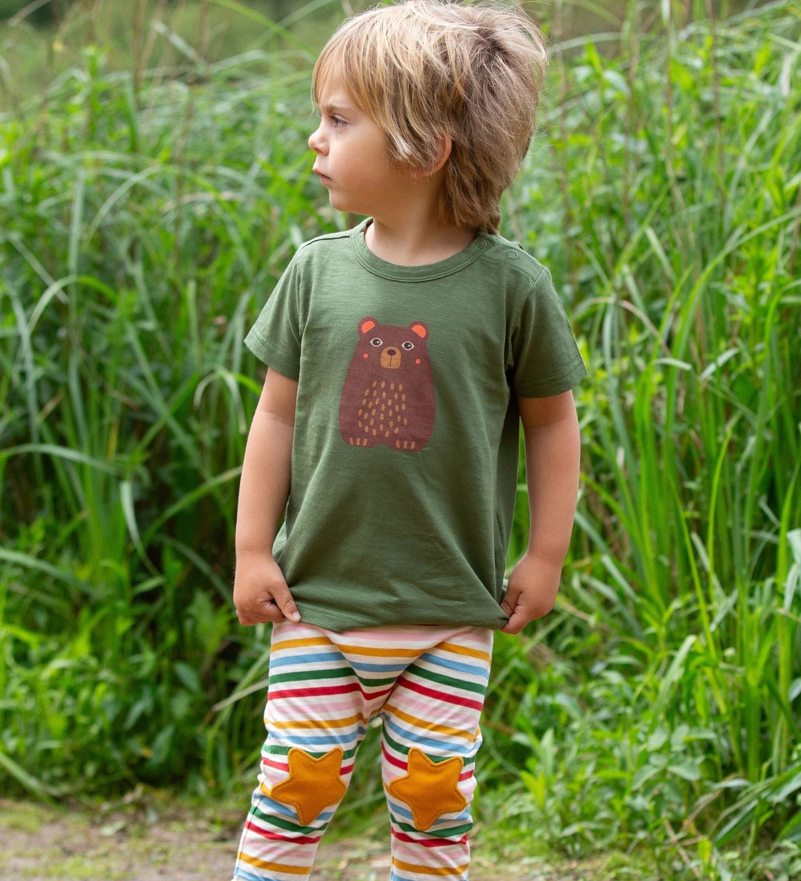 A child wearing the Little Green Radicals Kids Organic Cotton Green Bear Short Sleeve T-Shirt with rainbow striped star knee patch joggers.