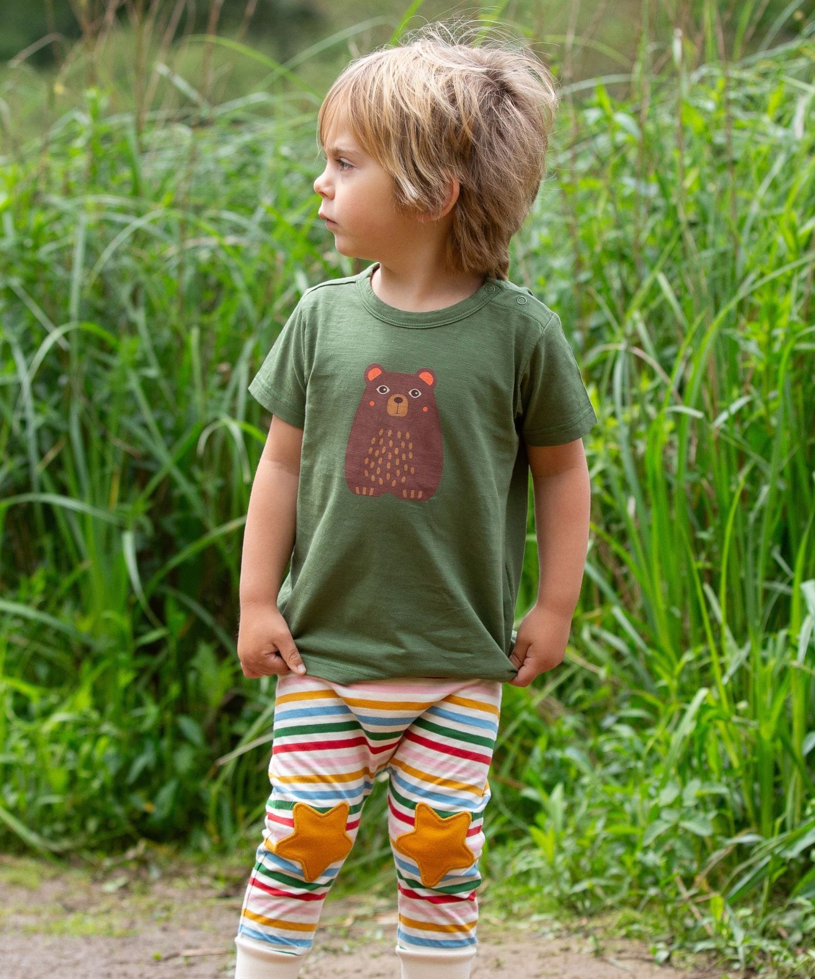 A child wearing the Little Green Radicals Kids Organic Cotton Green Bear Short Sleeve T-Shirt with rainbow striped star knee patch joggers.