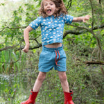 A child wearing the Little Green Radicals Kids Organic Cotton Blue Clouds Loose Fit Short Sleeve Henley Top with a pair of striped blue shorts. The child is jumping and has a pair of red wellies on.