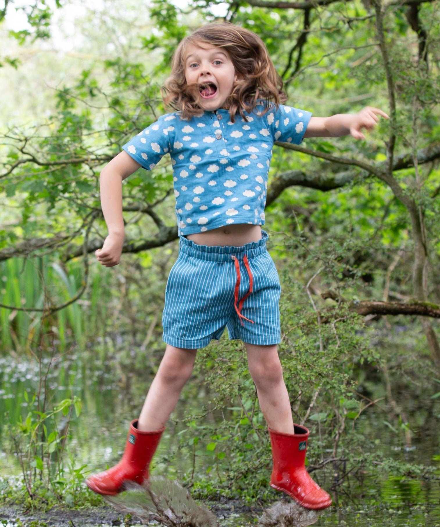 A child wearing the Little Green Radicals Kids Organic Cotton Blue Clouds Loose Fit Short Sleeve Henley Top with a pair of striped blue shorts. The child is jumping and has a pair of red wellies on.