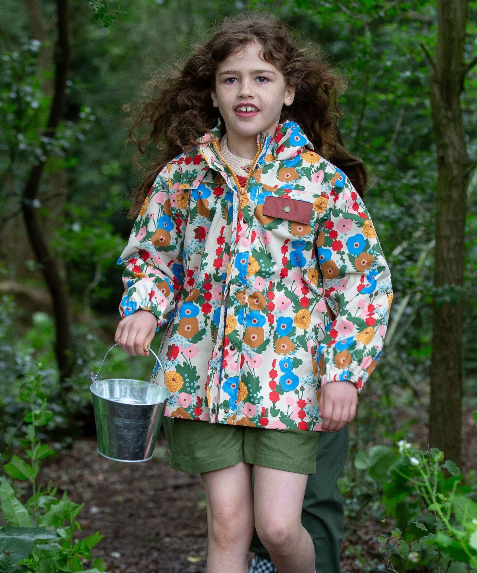 A child wearing the Little Green Radicals Child's Flora & Fauna vintage style flower print Waterproof Raincoat with green coloured shorts