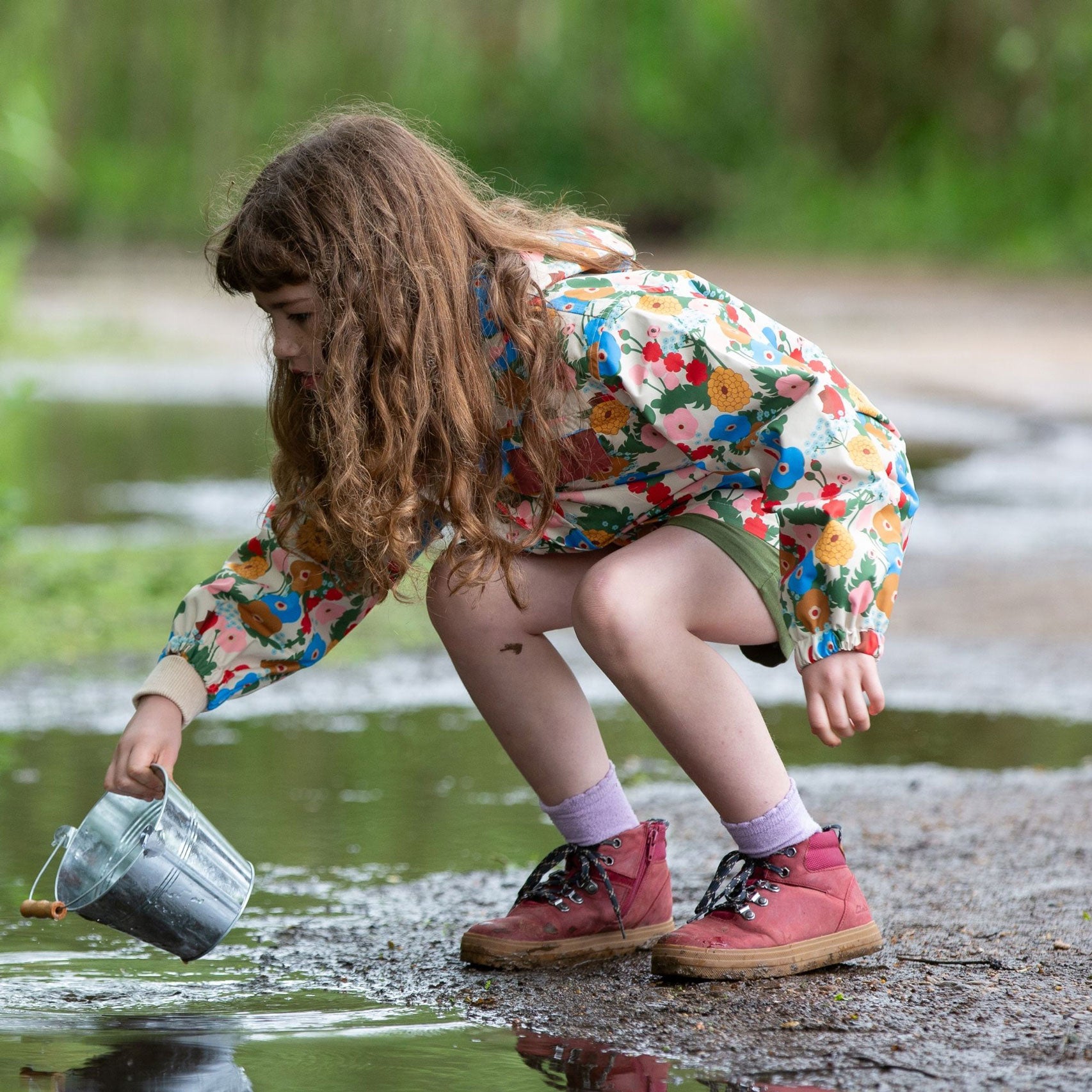 A child wearing the Little Green Radicals Child's Flora & Fauna vintage style flower print Waterproof Raincoat with green shorts and pink walking boots. The child is crouching down near water with a metal bucket in their hannd.