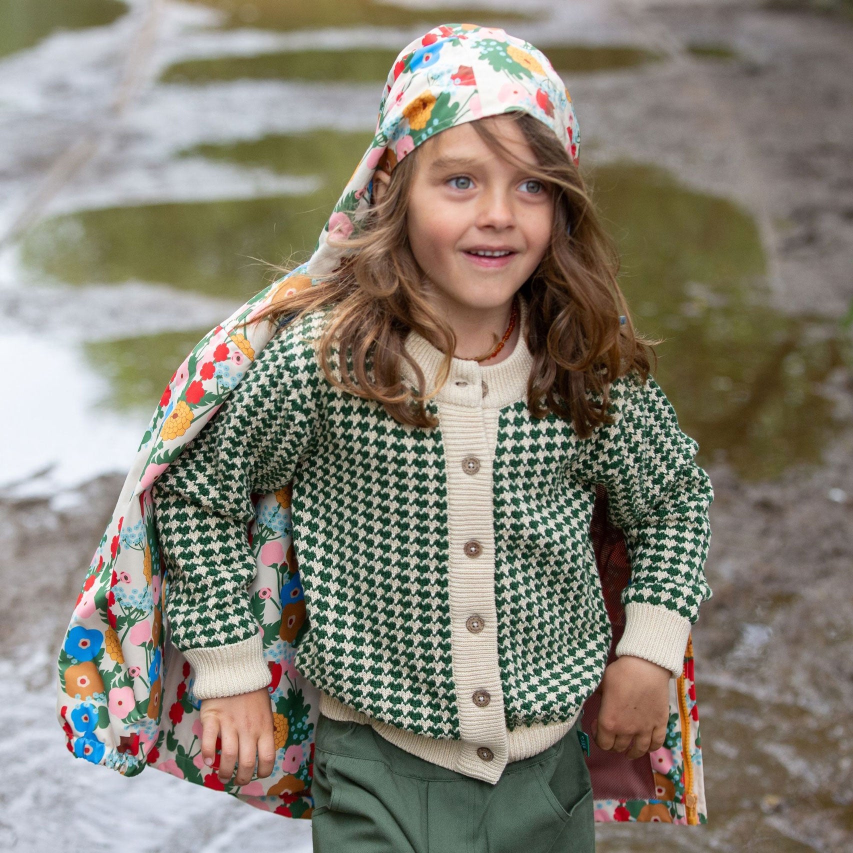 A child wearing the Little Green Radicals From One To Another Olive Green Striped Knitted Child's Organic Cotton Cardigan with green coloured shorts. The child has a floral print coat worn with just the hood on their head.