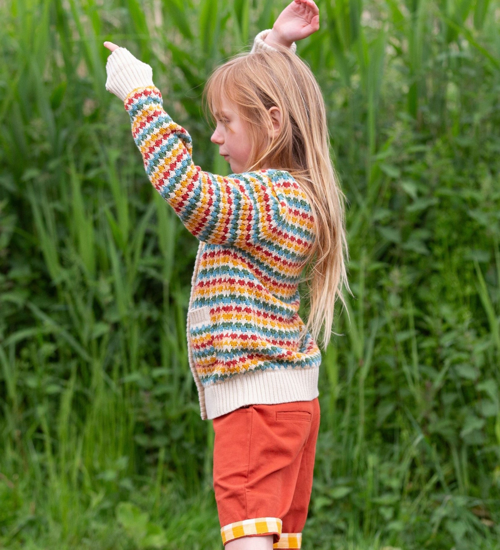 A child wearing the Little Green Radicals From One To Another Rainbow Days Organic Cotton Knitted Cardigan with red coloured shorts.