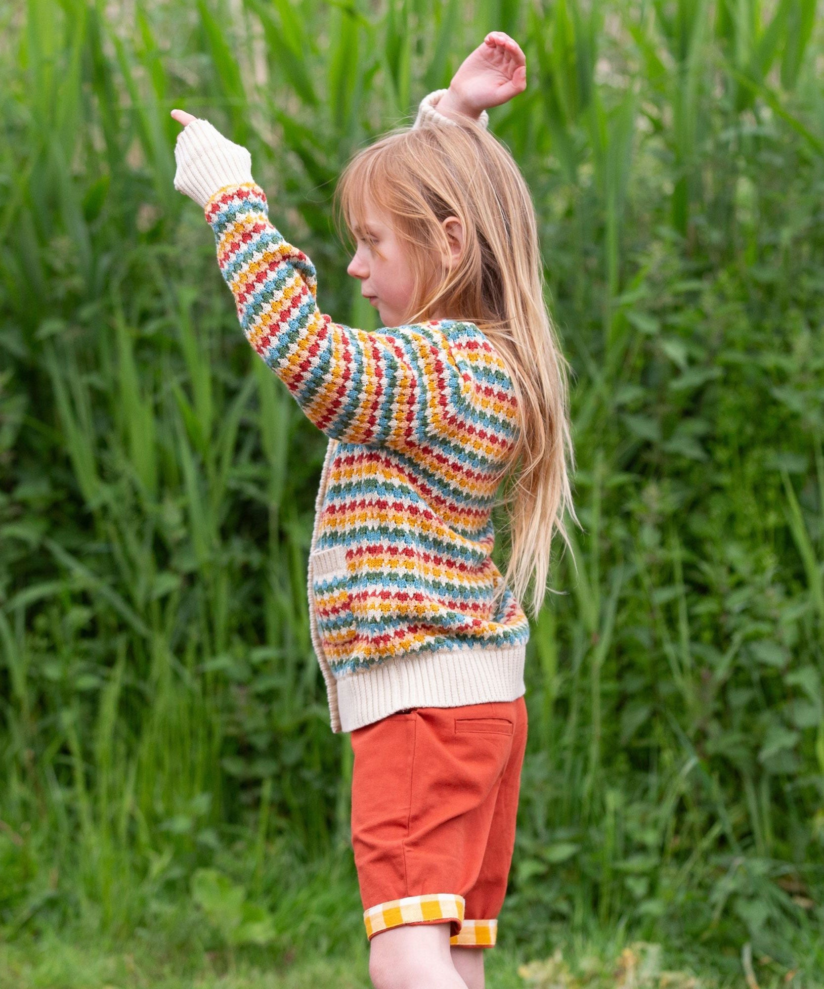 A child wearing the Little Green Radicals From One To Another Rainbow Days Organic Cotton Knitted Cardigan with red coloured shorts.