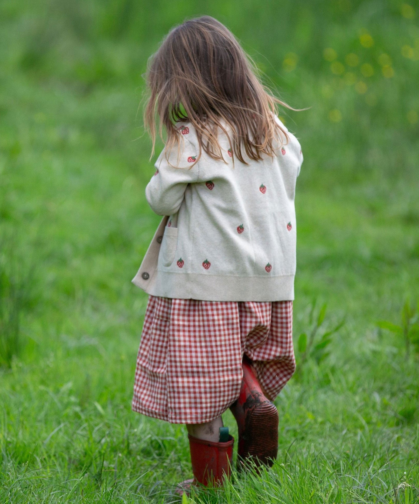 A child wearing the Little Green Radicals organic From One To Another Strawberry Days print child's Knitted Cardigan. The child is facing away from the camera showing the back of the cardigan