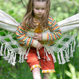 A child leaning on a hammock wearing the Little Green Radicals From One To Another Rainbow Days Organic Cotton Knitted Cardigan with red shorts.