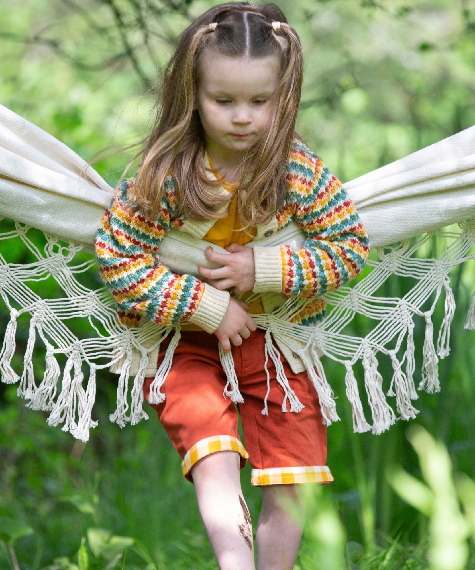 A child leaning on a hammock wearing the Little Green Radicals From One To Another Rainbow Days Organic Cotton Knitted Cardigan with red shorts.