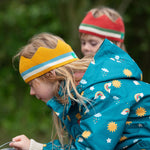 A child wearing the Little Green Radicals Child's Rainbow Days print Recycled Waterproof Raincoat with a yellow gold coloured striped knitted crown. 