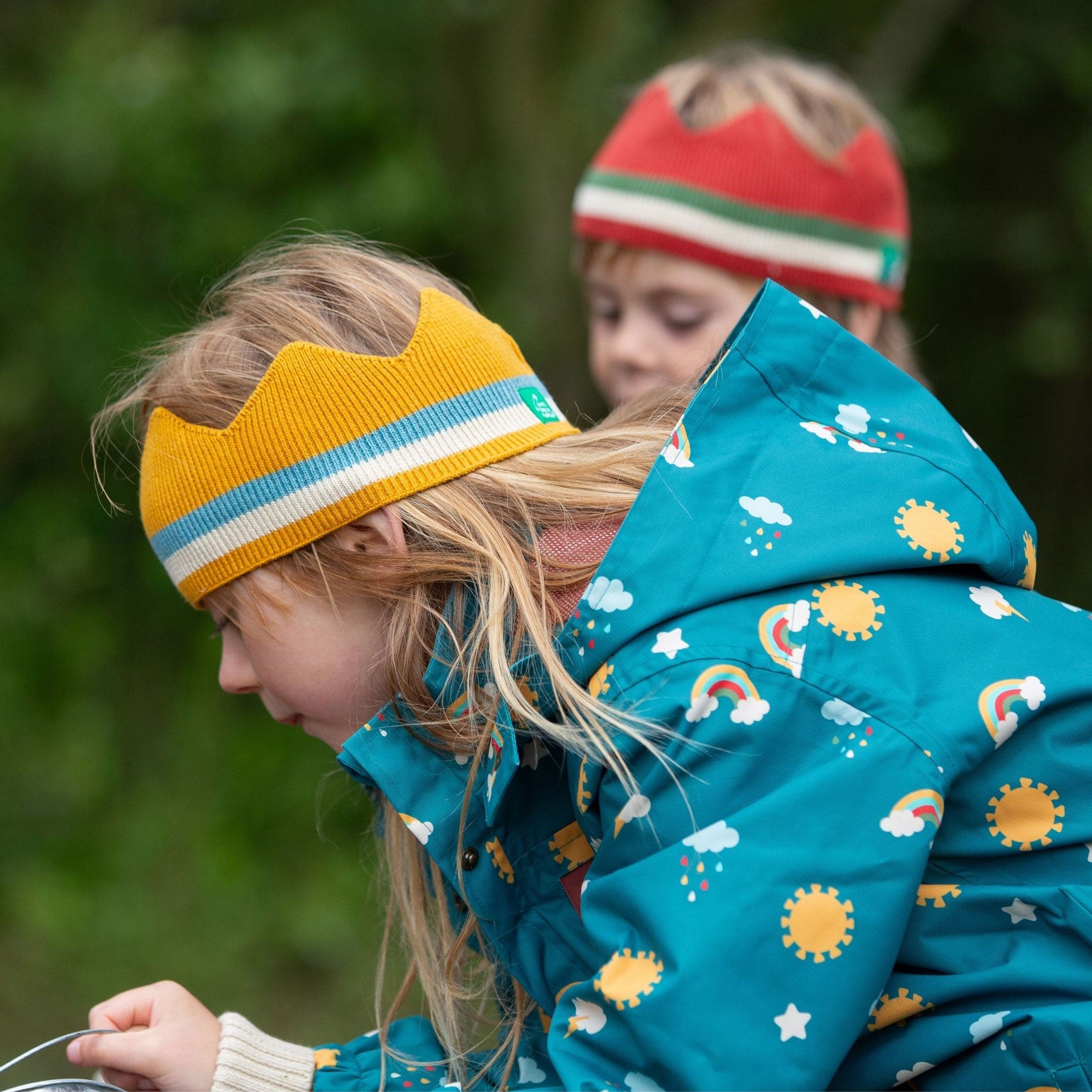 A child wearing the Little Green Radicals Child's Rainbow Days print Recycled Waterproof Raincoat with a yellow gold coloured striped knitted crown. 