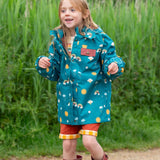 A child wearing the Little Green Radicals Child's Rainbow Days print Recycled Waterproof Raincoat with red rust coloured shorts and red wellington boots. 