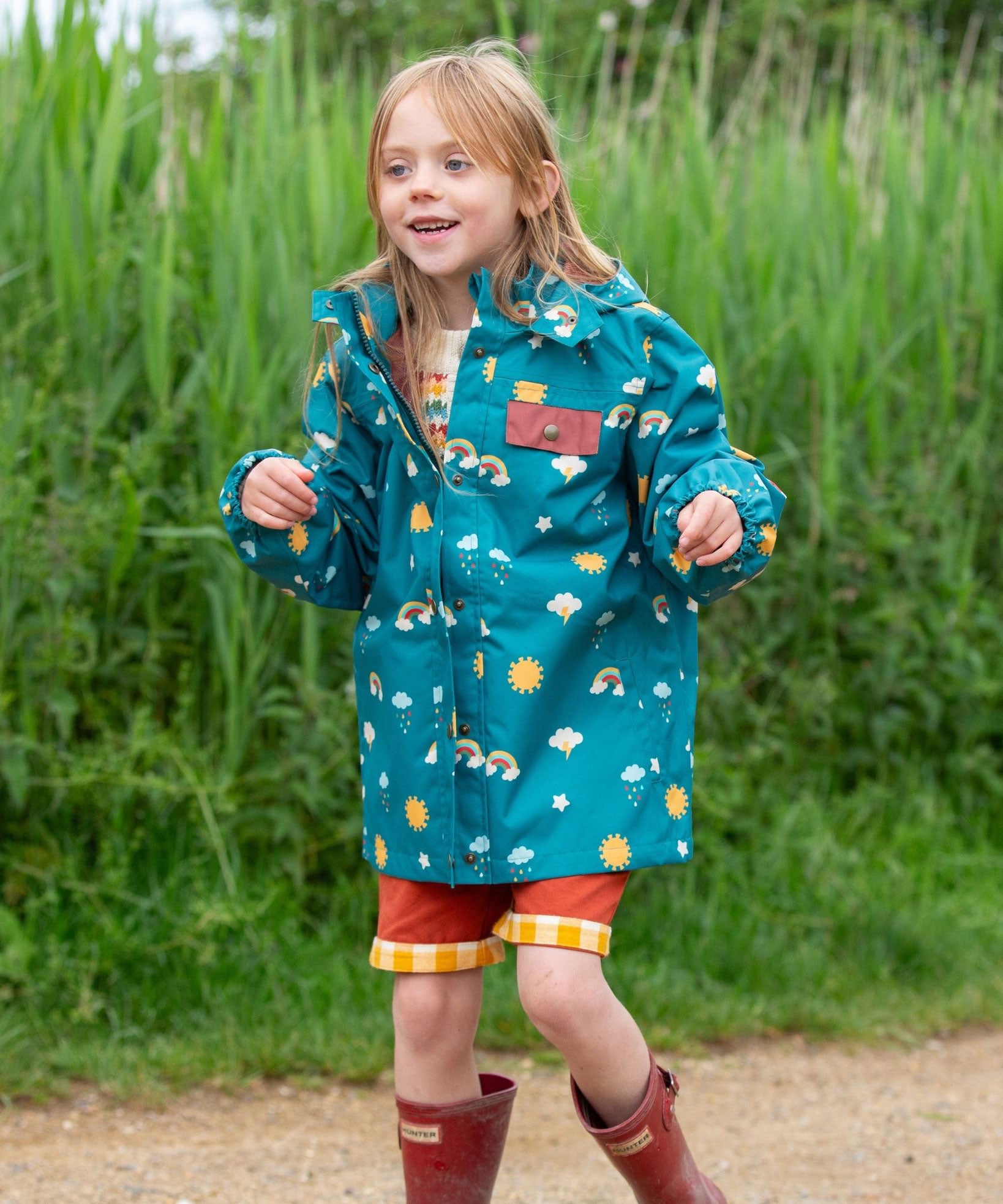 A child wearing the Little Green Radicals Child's Rainbow Days print Recycled Waterproof Raincoat with red rust coloured shorts and red wellington boots. 