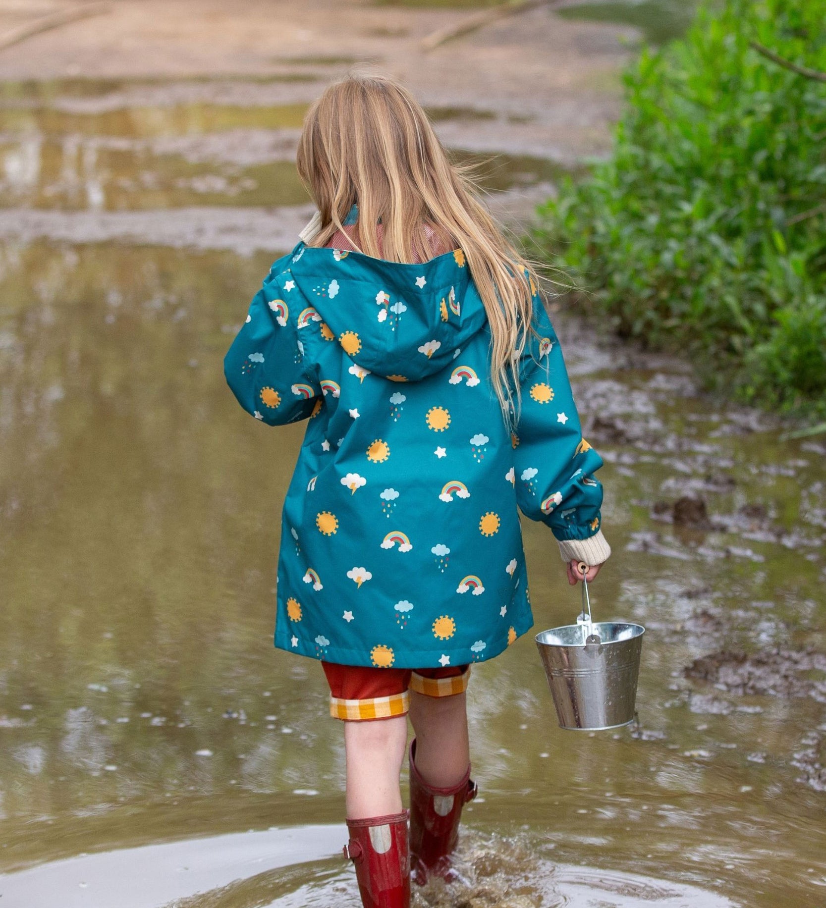 A child wearing the Little Green Radicals Child's Rainbow Days print Recycled Waterproof Raincoat facing away from the camera showing the back of the jacket