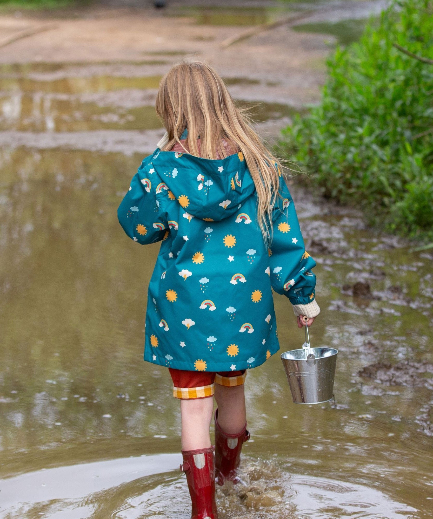 A child wearing the Little Green Radicals Child's Rainbow Days print Recycled Waterproof Raincoat facing away from the camera showing the back of the jacket