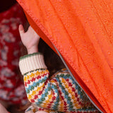 A child sitting in an orange coloured tent, the child is wearing the Little Green Radicals From One To Another Rainbow Days Organic Cotton Knitted Cardigan, the child's arm can be seen peeping out the side of the tent.