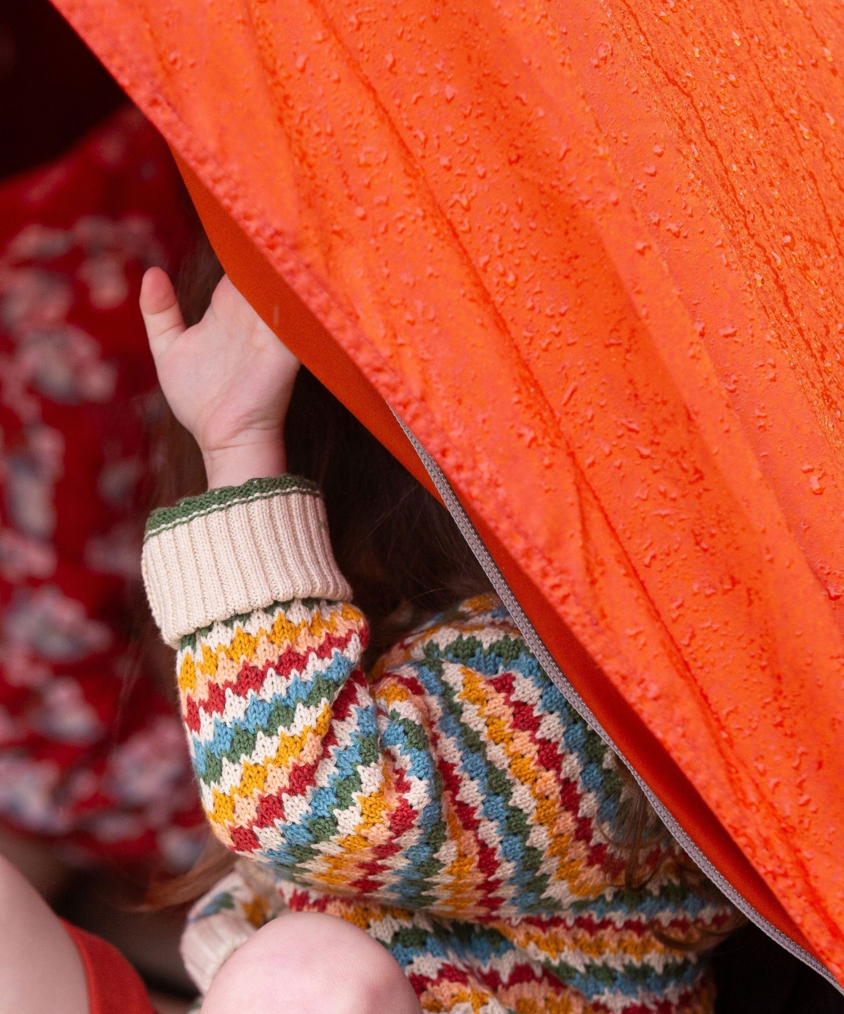 A child sitting in an orange coloured tent, the child is wearing the Little Green Radicals From One To Another Rainbow Days Organic Cotton Knitted Cardigan, the child's arm can be seen peeping out the side of the tent.