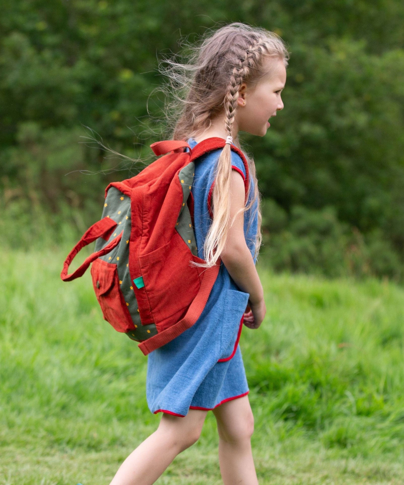 A side view of a child wearing the Little Green Radicals Kids Ready For Adventure Yellow Stars print red and green Backpack on their back showing the side of the bag. 