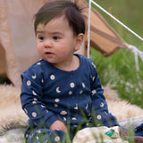 A baby wearing the Little Green Radicals Full Moon Organic Cotton Babygrow sitting in front of a tent outdoors. 