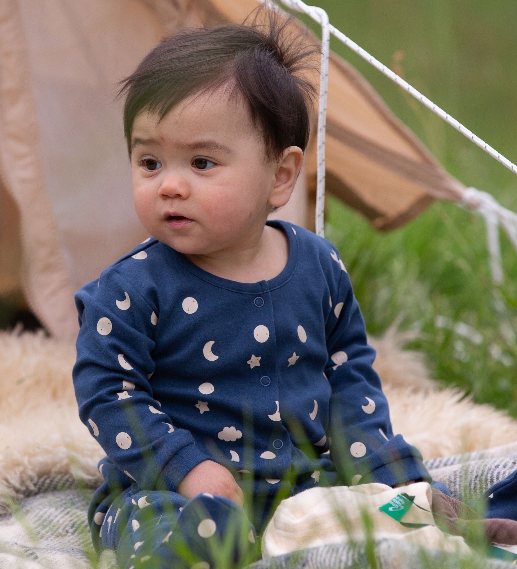 A baby wearing the Little Green Radicals Full Moon Organic Cotton Babygrow sitting in front of a tent outdoors. 