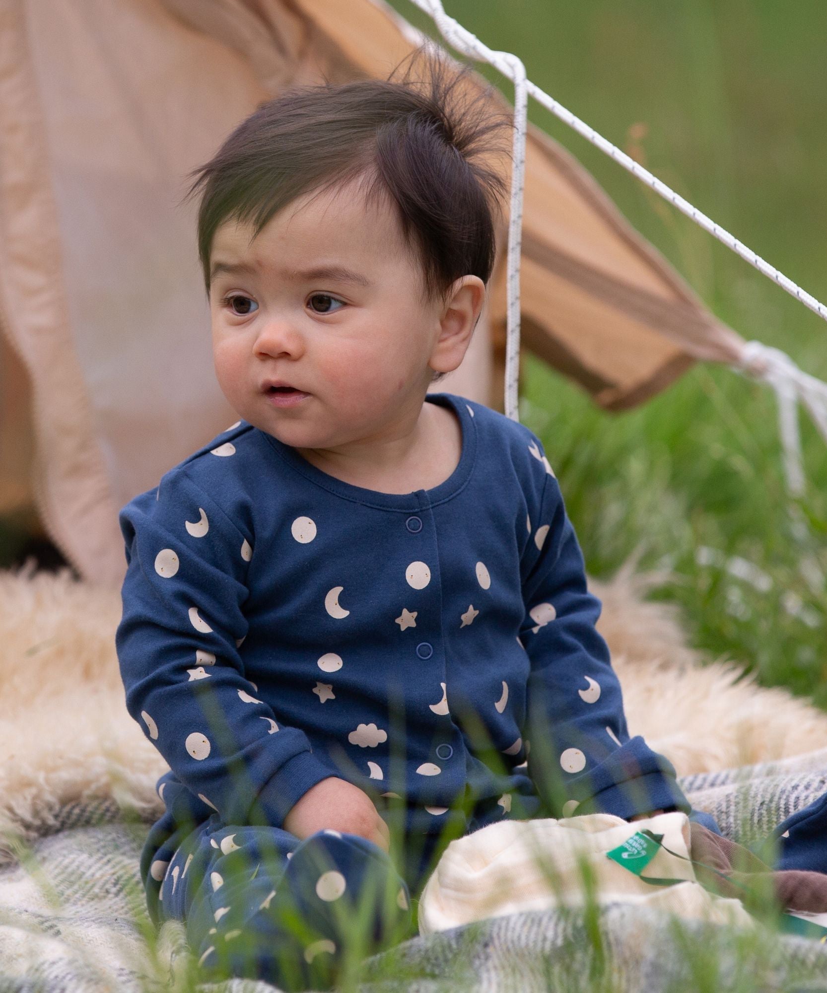 A baby wearing the Little Green Radicals Full Moon Organic Cotton Babygrow sitting in front of a tent outdoors. 